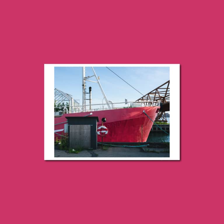 Postkarte vor pinkem Hintergrund: der rote Rumpf eines Schiffs am Kai von Refshaleøen in Kopenhagen, davor eine dunkle Hütte.