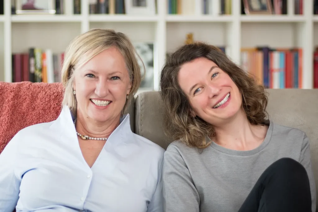 Dr. med. Bärbel Grashoff und Marie Luisa Kerkhoff sitzen lachend nebeneinander auf einem Sofa vor einem Bücherregal, Bärbel Grashoff hat eine Hand auf Marie Luisa Kerkhoffs Schulter.