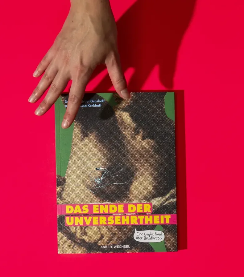 Eine Hand legt das Buch „Das Ende der Unversehrtheit“ auf einen pinken Untergrund, das Cover zeigt einen gerasterten weiblichen Oberkörper mit gezeichnetem Sprung auf der Brust.