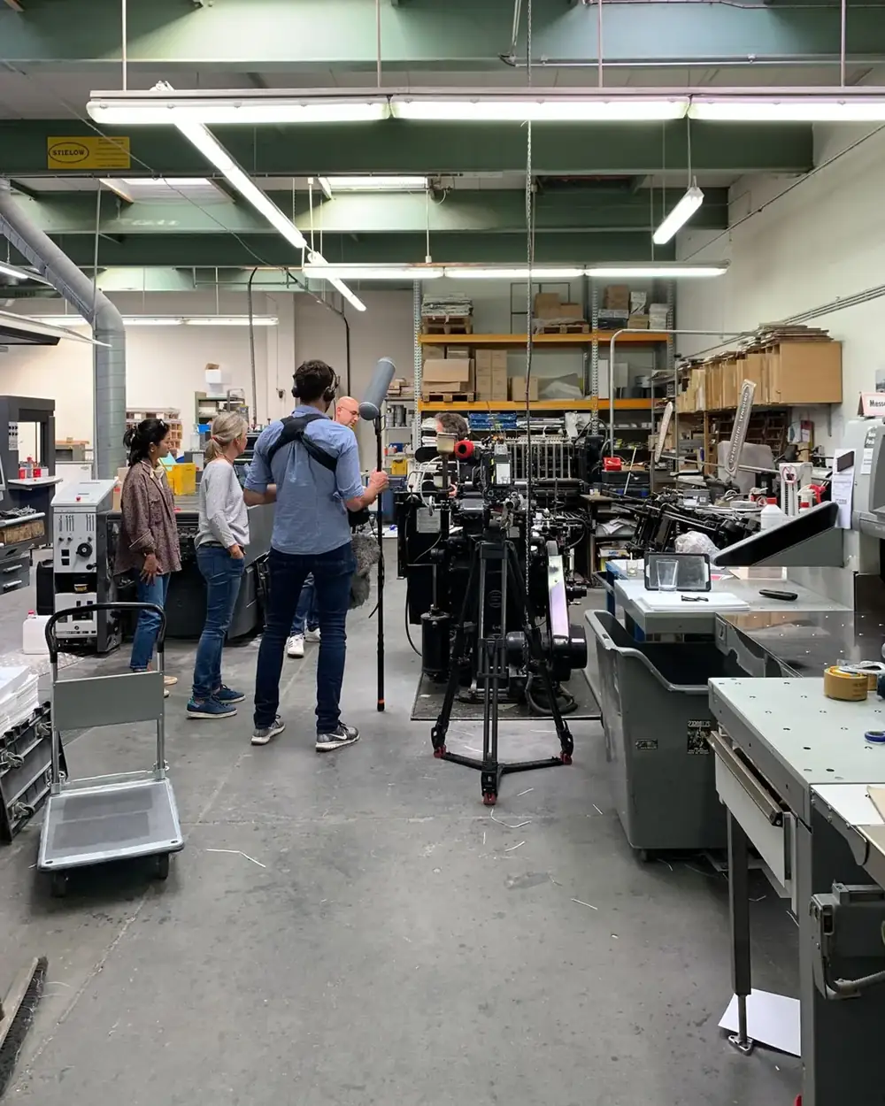 Eine Druckerei mit Maschinen und Papierstapeln, ein Kamerateam mit Mikrofonangel filmt an einer Druckmaschine