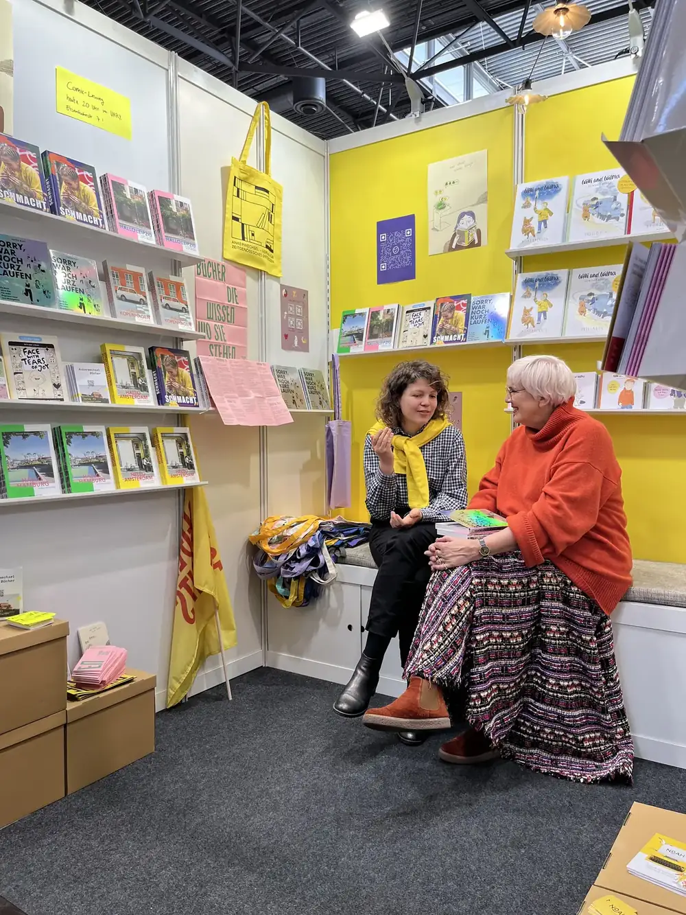 Harriet Dohmeyer im Gespräch mit Claudia Roth am Messestand des Ankerwechsel Verlags, ringsum Bücher in gelben Regalen
