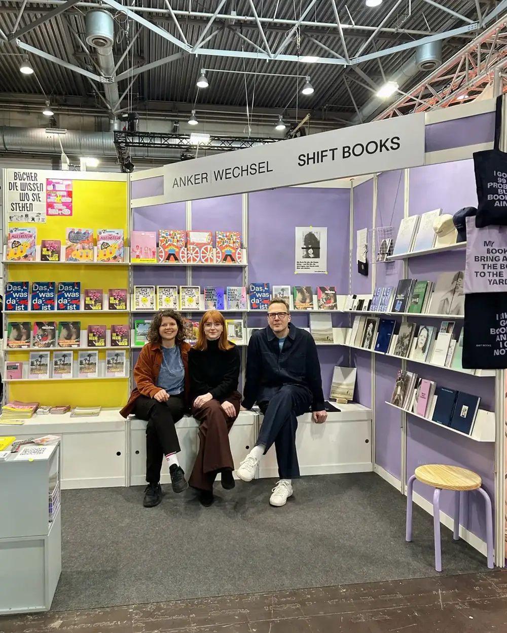 Ein Messestand mit der Beschriftung Anker Wechsel und Shift Books, Bücher in gelben und lila Regalen, drei Menschen sitzen auf der Bank davor