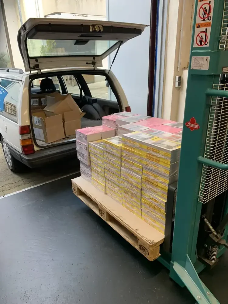 Eine Palette mit frisch gedruckten Büchern auf einem Hubwagen, daneben ein Kombi mit offener Heckklappe und Versandkartons